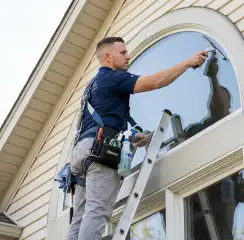 Professional window cleaner on a ladder using a squeegee to clean an exterior arched window, Denver, CO.